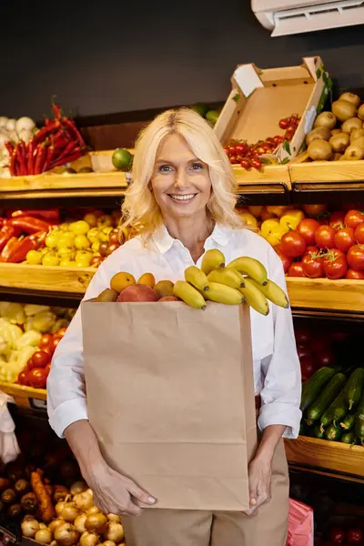 Good looking mature woman in casual attire holding shopping bag full of fruits and smiling happily — Stock Photo