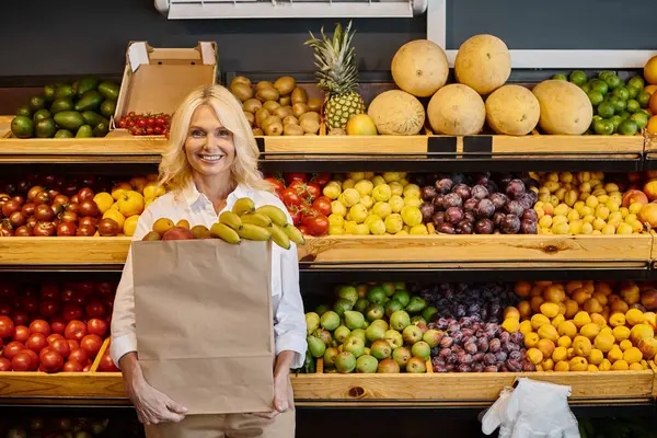 Alegre atractiva mujer madura en ropa cotidiana sonriendo y sosteniendo bolsa de compras con plátanos - foto de stock