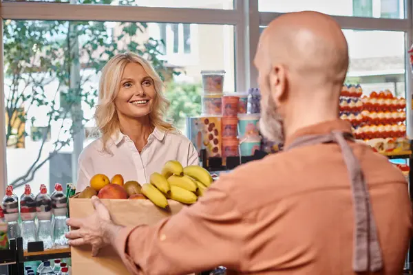 Сосредоточьтесь на веселой зрелой клиентке улыбаясь размытому продавцу с сумкой, полной фруктов — стоковое фото