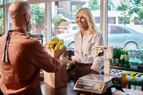 Focus on jolly mature customer smiling at blurred seller next to cash desk at grocery store — Stock Photo