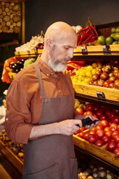 Vendedor barbudo maduro mirando etiquetas de precios en sus manos con puesto de comestibles con frutas en el telón de fondo - foto de stock
