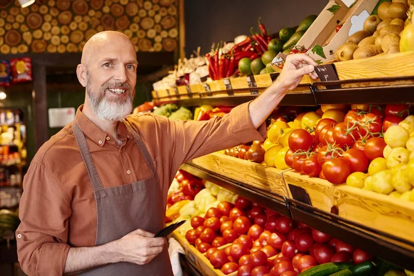 Бодрый бородатый продавец наклеивает ценники на свежие яркие продукты и радостно улыбается в камеру — стоковое фото