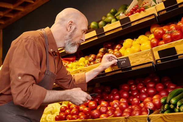 Nachdenklicher Verkäufer mit grauem Bart klebt Preisschilder an frischem Gemüse im Supermarkt — Stockfoto
