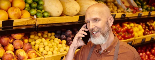 Gray bearded mature salesman talking cheerfully by phone with fruits and vegetables on background — Stock Photo