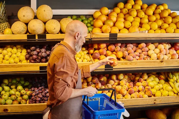 Grauer bärtiger, gut aussehender Verkäufer, der frischen Pfirsich in der Hand hält und einen Einkaufskorb hält — Stockfoto