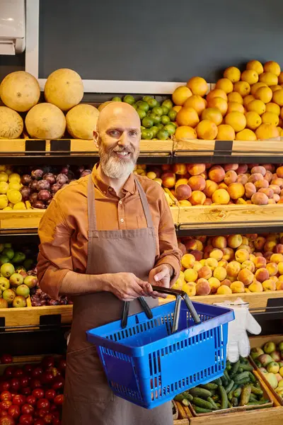 Cheerful mature seller with shopping basket in hands smiling at camera with fruits on backdrop — Stock Photo