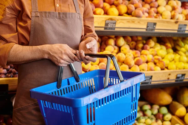 Cropped view of mature salesman holding shopping basket in hands with blurred fruits on background — Stock Photo