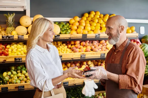 Fröhlicher bärtiger Verkäufer, der seinem fröhlichen Kunden eine Packung Erdbeeren schenkt, einander lächelnd — Stockfoto