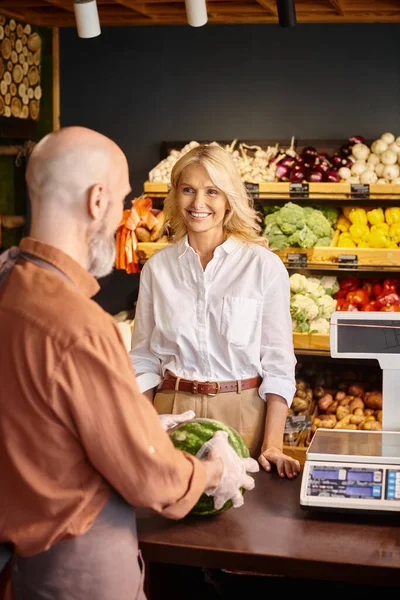 Fokus auf fröhliche reife Frau lächelt verschwommenen Verkäufer mit Bart wiegt frische riesige Wassermelone — Stockfoto