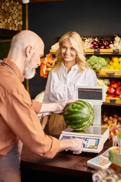 Focus on mature female customer smiling at blurred bearded seller weighing fresh watermelon — Stock Photo