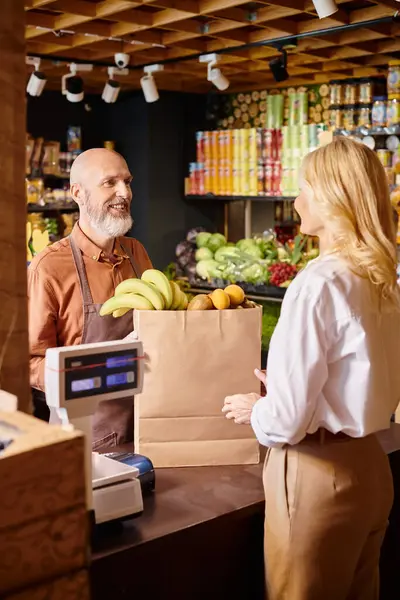 Зрілий веселий продавець з бородою посміхається своєму клієнту, що купує свіжі фрукти в продуктовому магазині — стокове фото