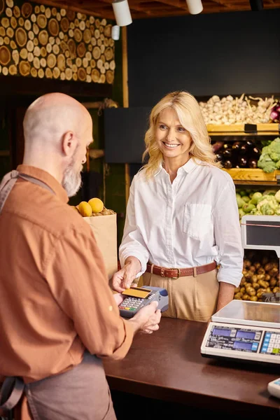 Focus on joyful mature customer paying with her credit card and smiling at blurred bearded seller — Stock Photo