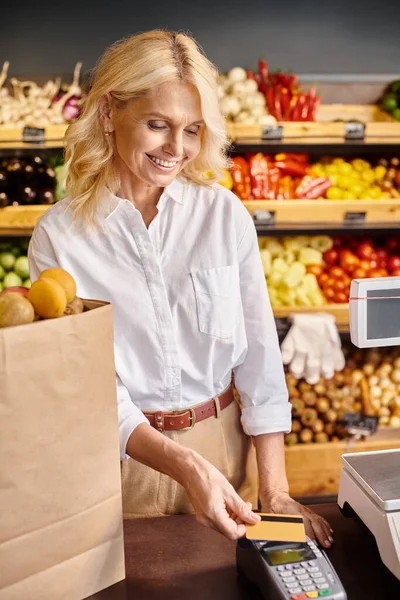 Maduro feliz cliente femenino con bolsa de compras con frutas en el mostrador de efectivo que paga con su tarjeta de crédito - foto de stock
