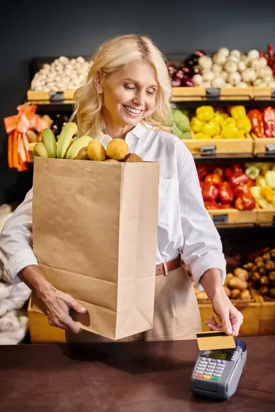 Alegre mujer madura en traje casual sosteniendo bolsa de compras y pagando con su tarjeta de crédito - foto de stock