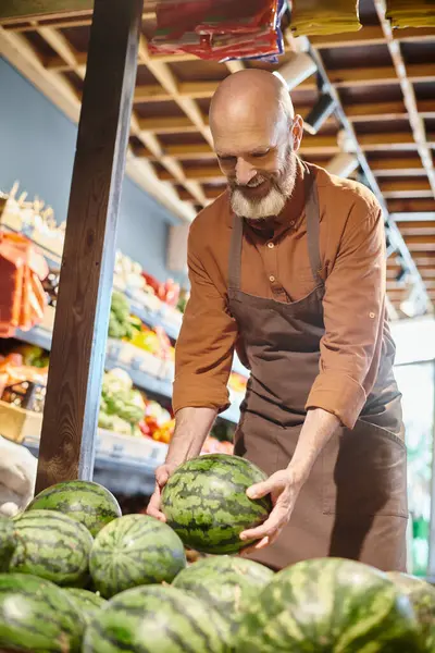 Веселый бородатый зрелый продавец, счастливо улыбающийся и собирающий свежий арбуз в продуктовом магазине — стоковое фото
