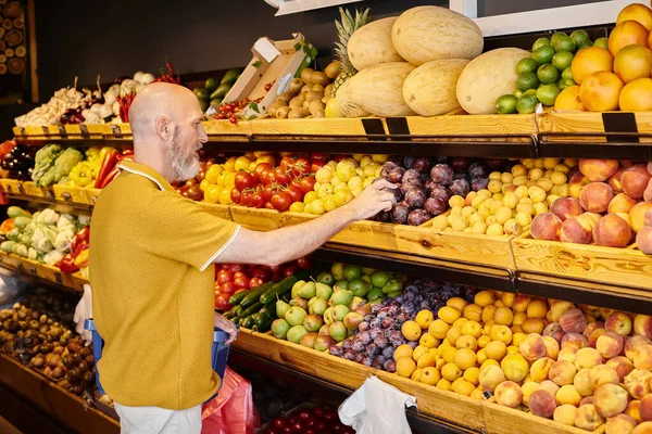 Bearded male customer in casual attire with shopping basket choosing fresh plums at grocery store — Stock Photo