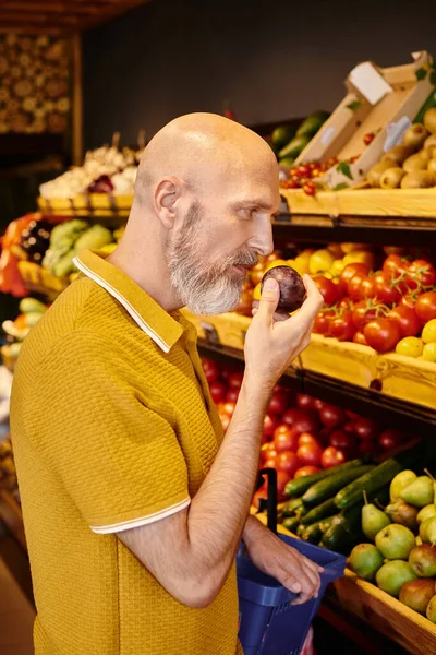 Bärtiger älterer Mann in Freizeitkleidung mit Einkaufskorb und frischen Pflaumen im Supermarkt — Stockfoto
