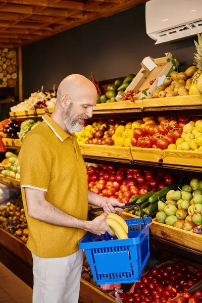 Веселий зрілий клієнт у повсякденному вбранні вставляє свіжі яскраві банани в кошик для покупок — стокове фото