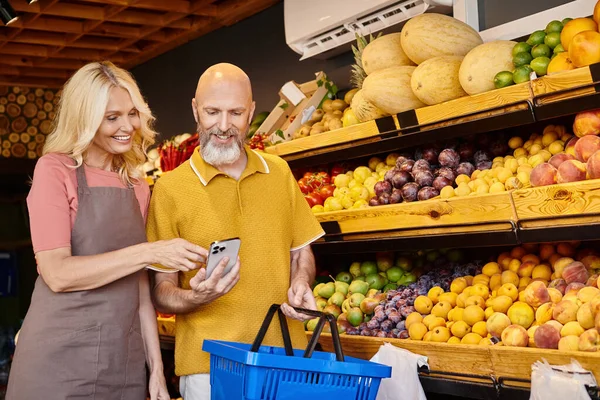 Reife Verkäuferin hilft graubärtigem Mann bei der Auswahl von Lebensmitteln und beide schauen aufs Handy — Stockfoto