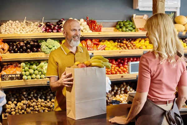 Centrarse en el cliente maduro comprar frutas frescas y sonreír al vendedor femenino borroso en la tienda de comestibles - foto de stock