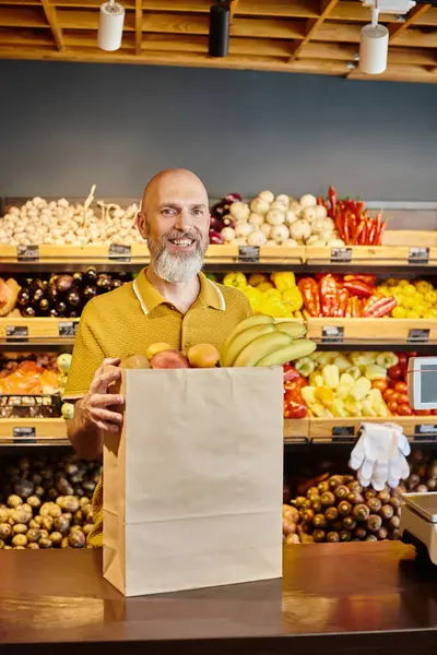 Hombre barbudo alegre posando con bolsa de papel llena de frutas frescas y sonriendo a la cámara en el supermercado - foto de stock