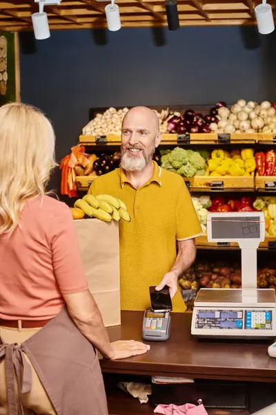 Fokus auf lustigen bärtigen Mann, der mit seinem Handy für frische Lebensmittel bezahlt und Verkäuferin anlächelt — Stockfoto