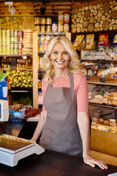 Fröhliche reife Verkäuferin posiert an der Kasse und lächelt im Supermarkt fröhlich in die Kamera — Stockfoto