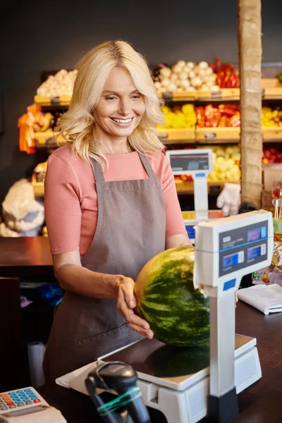 Freudige reife Verkäuferin wiegt saftige Wassermelone an der Kasse und lächelt glücklich im Supermarkt — Stockfoto