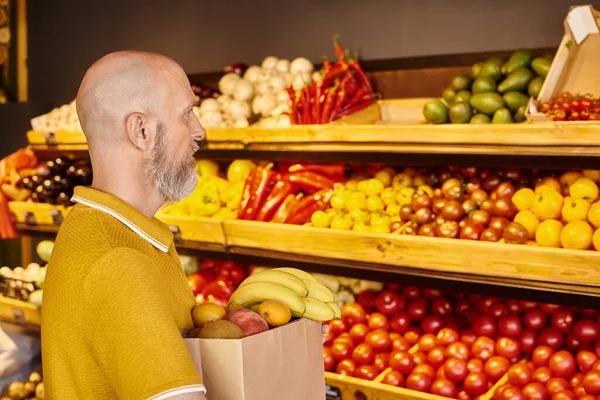 Hombre barbudo gris maduro concentrado sosteniendo bolsa de papel llena de frutas frescas en la tienda de comestibles - foto de stock