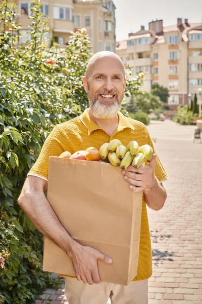 Fröhliche reife männliche Kundin posiert draußen mit Papiertüte voller Früchte und lächelt in die Kamera — Stockfoto