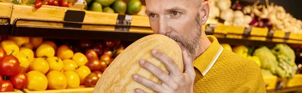 Konzentrierte graubärtige männliche Kundin schnüffelt lebhafte nahrhafte Melone im Lebensmittelgeschäft, Banner — Stockfoto