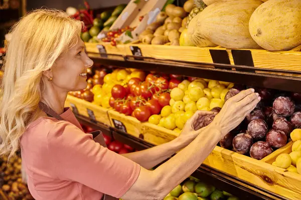 Commessa jolly maturo mettendo prugne fresche e vivaci su bancarella con altri frutti al negozio di alimentari — Foto stock
