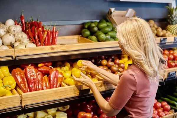 Vendedora rubia madura sosteniendo tomates amarillos frescos junto a la tienda de verduras en la tienda de comestibles - foto de stock