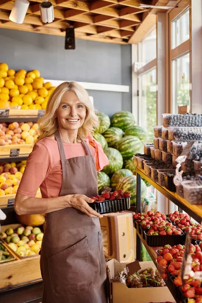 Fröhliche reife Verkäuferin, die köstliche frische Erdbeeren in der Hand hält und glücklich in die Kamera lächelt — Stockfoto
