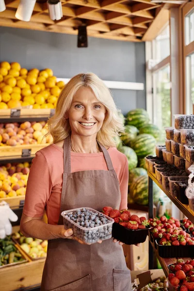Joyful mature saleswoman holding fresh vibrant strawberries and blueberries and smiling at camera — Stock Photo