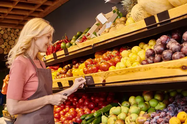 Mature attractive jolly saleswoman putting price tags on vegetable stall and smiling happily — Stock Photo