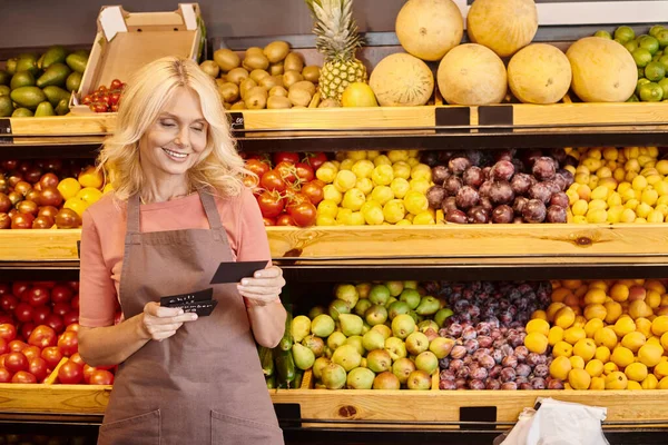 Happy blonde mature saleswoman looking at price tags with vegetables and fruits on background — Stock Photo