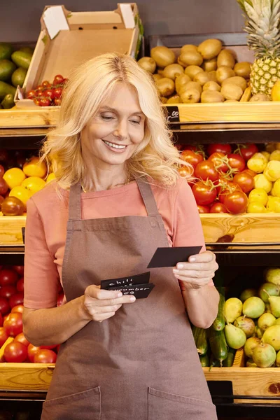 Vertikale Aufnahme einer reifen, fröhlichen Verkäuferin, die lächelt und auf Preisschilder im Lebensmittelgeschäft blickt — Stockfoto