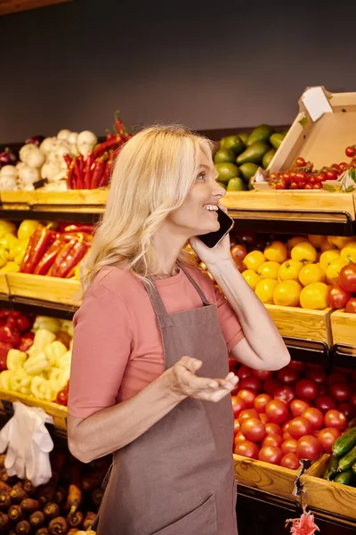 Attraktive, gut gelaunte Verkäuferin, die mit dem Handy telefoniert und frisches Obst und Gemüse betrachtet — Stockfoto