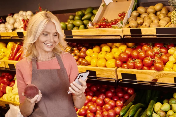 Pretty mature saleswoman looking happily at her mobile phone and holding fresh nutritious plum — Stock Photo
