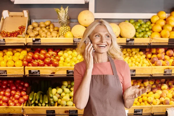 Fröhliche attraktive Verkäuferin lächelt und telefoniert mit Obst und Gemüse vor dem Hintergrund — Stockfoto