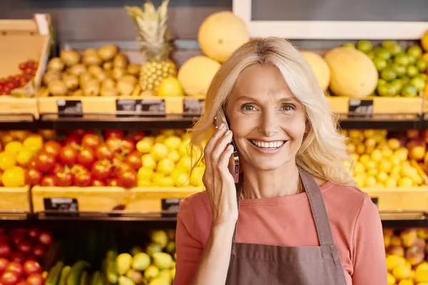 Joyful mature blonde saleswoman talking by mobile phone and smiling at camera at grocery store — Stock Photo