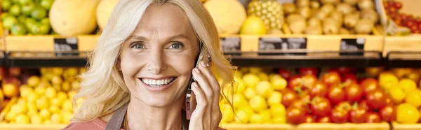 Cheerful mature saleswoman talking by mobile phone and smiling at camera at grocery shop, banner — Stock Photo