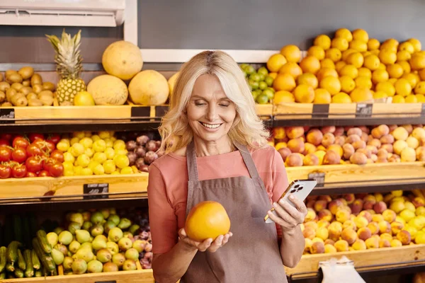 Attraktive lustige Verkäufer halten Handy und frische nahrhafte riesige Orange und lächeln glücklich — Stockfoto