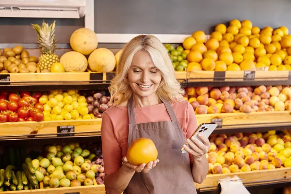 Atractivo vendedor alegre celebración de teléfono móvil y fresco nutritivo enorme naranja y sonriendo felizmente - foto de stock