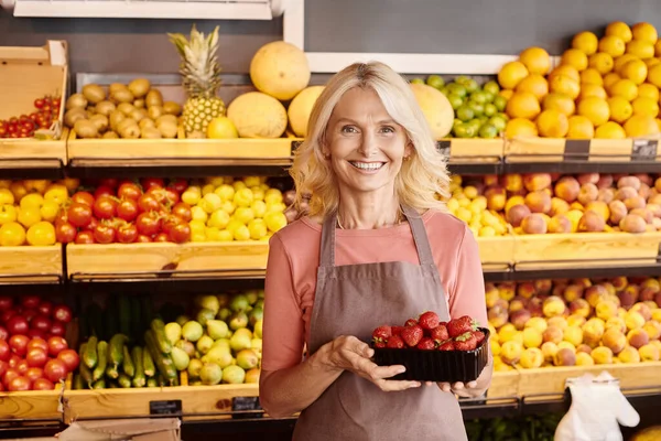Glückliche reife Verkäuferin posiert mit leckeren Erdbeeren in den Händen und lächelt fröhlich in die Kamera — Stockfoto