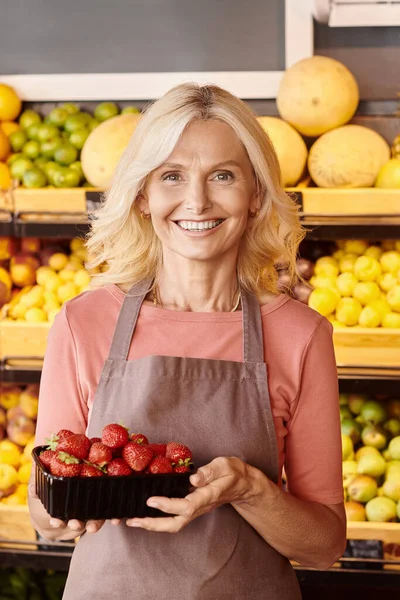 Tiro vertical de alegre vendedor maduro sosteniendo paquete de fresas frescas nutritivas sonriendo felizmente - foto de stock