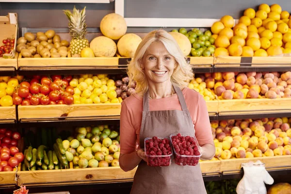 Alegre vendedora madura sosteniendo dos paquetes de frescas frambuesas vibrantes y sonriendo a la cámara - foto de stock
