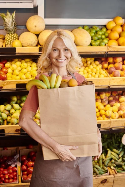 Alegre madura vendedora sosteniendo bolsa de papel llena de frutas frescas y sonriendo a la cámara en la tienda - foto de stock