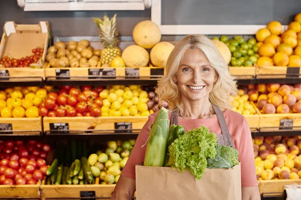 Fröhliche reife Verkäuferin mit Papiertüte voller Grünzeug in der Hand, die im Supermarkt in die Kamera lächelt — Stockfoto